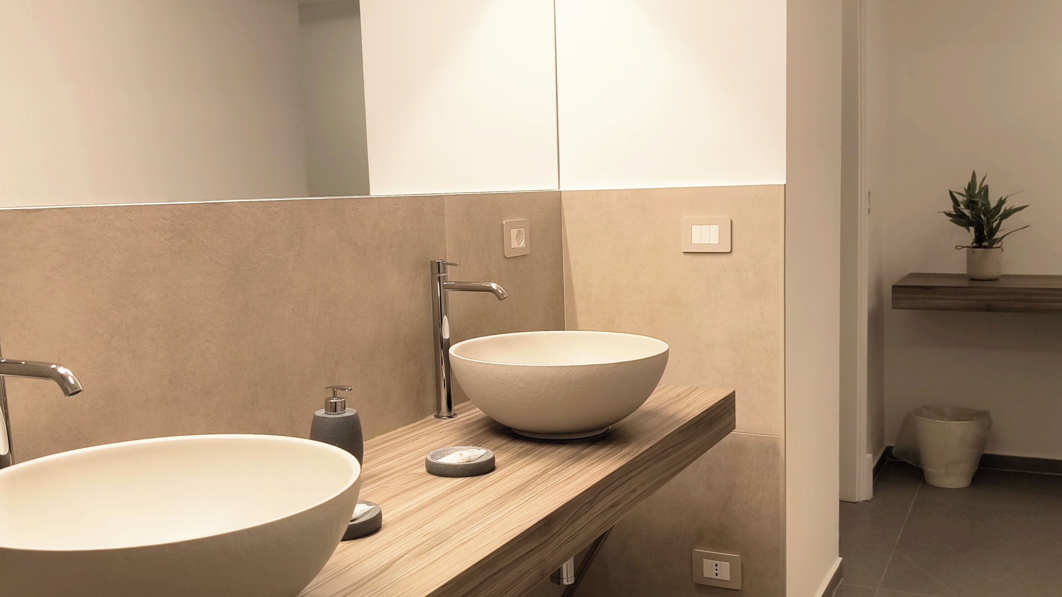 A modern bathroom with two large vessel sinks, a wooden vanity, and a small potted plant on a shelf.