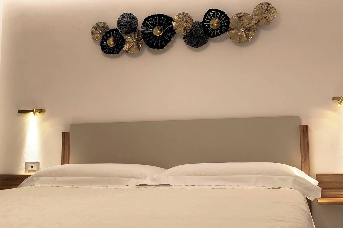 Quarto de hotel com uma cama, duas luzes fixadas na parede e um objeto decorativo na parede.