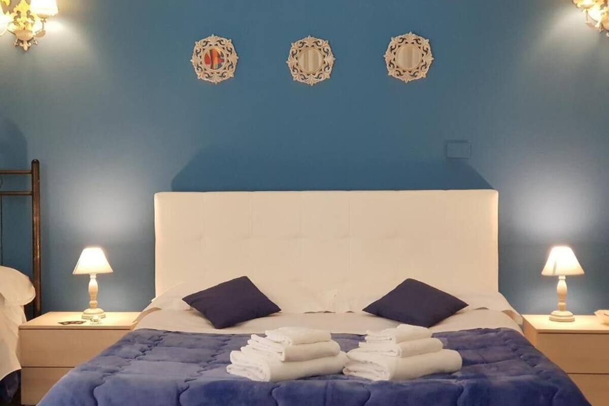 Quarto com parede azul, cabeceira branca, duas luminárias de cabeceira, cama com colcha azul, duas almofadas e três espelhos decorativos na parede.