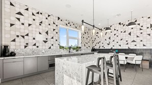 Et moderne køkken med flisebeklædt backsplash i geometriske mønstre, en stor ø med barstole og et spiseområde med et hvidt bord og stole.