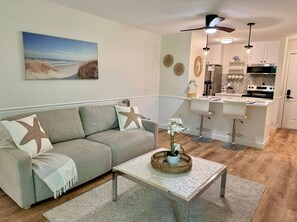 A living room with a sofa, coffee table, and a painting of a beach. - New Cape Cod Condo Near Beaches (Brewster)