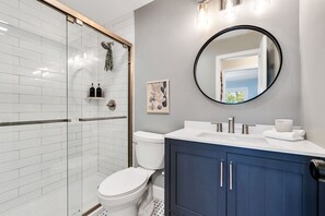 A modern bathroom with a glass shower enclosure, a white toilet, a vanity with a dark blue cabinet, and a round mirror.