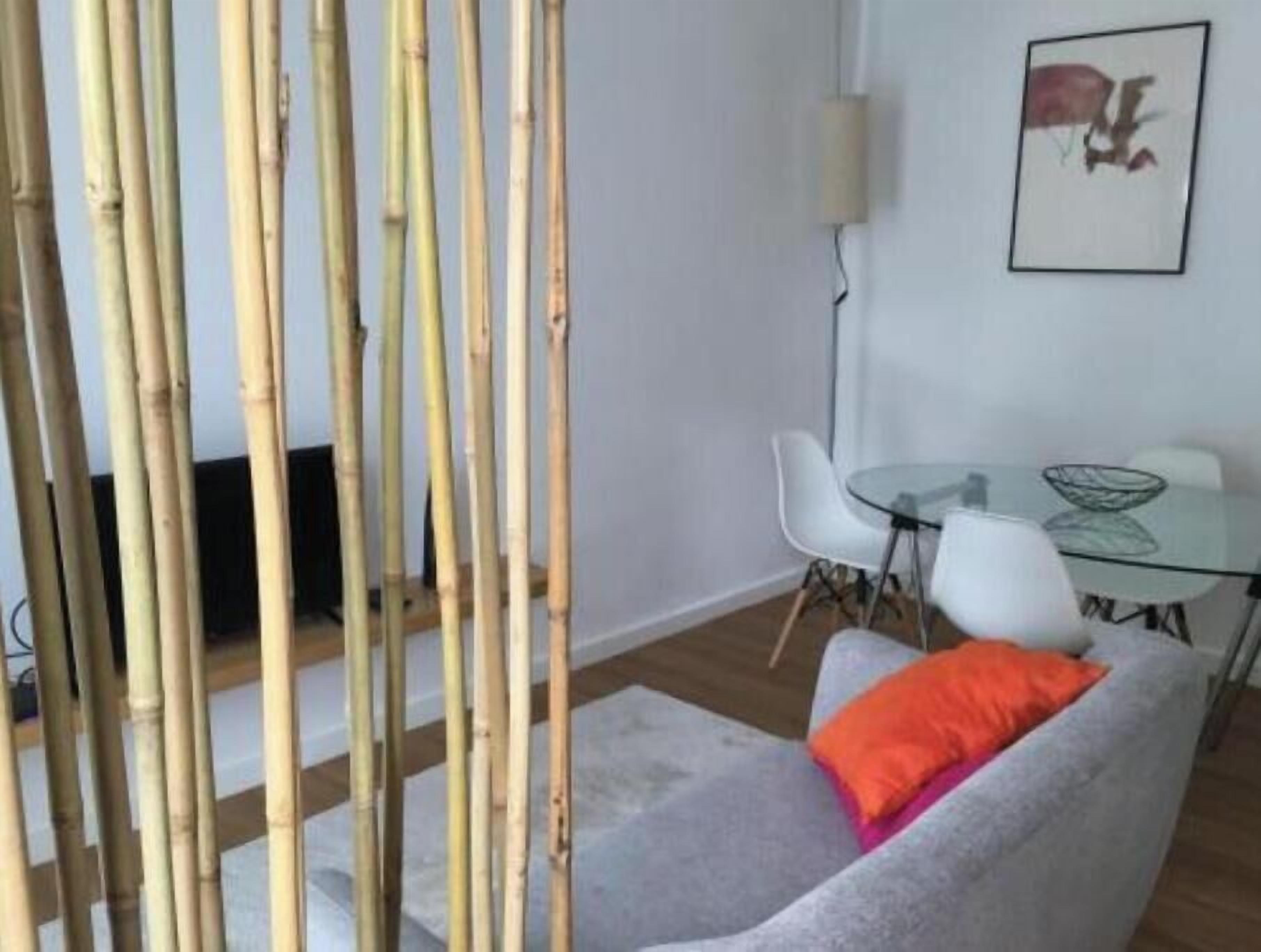 Une pièce avec une table en verre, des chaises blanches, un canapé gris agrémenté d’un coussin orange et des cloisons en bambou.