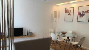Ein modernes Wohnzimmer mit einem Glastisch, weißen Stühlen, einer Couch, einem Flachbildfernseher und gerahmten Kunstwerken an der Wand.