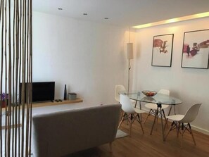 Ein modernes Wohnzimmer mit einem Glastisch, weißen Stühlen, einer Couch, einem Flachbildfernseher und gerahmten Kunstwerken an der Wand.