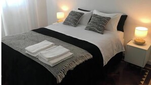 Ein ordentlich gemachtes Bett mit einer schwarz-weißen Tagesdecke, zwei Kissen und einem gefalteten weißen Handtuch auf einer grauen Decke. Auf den weißen Nachttischen stehen je eine Nachttischlampe.