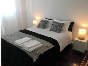 A neatly made bed with a black and white bedspread, two pillows, and a folded white towel on a gray blanket. There are two bedside lamps on white nightstands. - Pedra do Sal House (Estoril)
