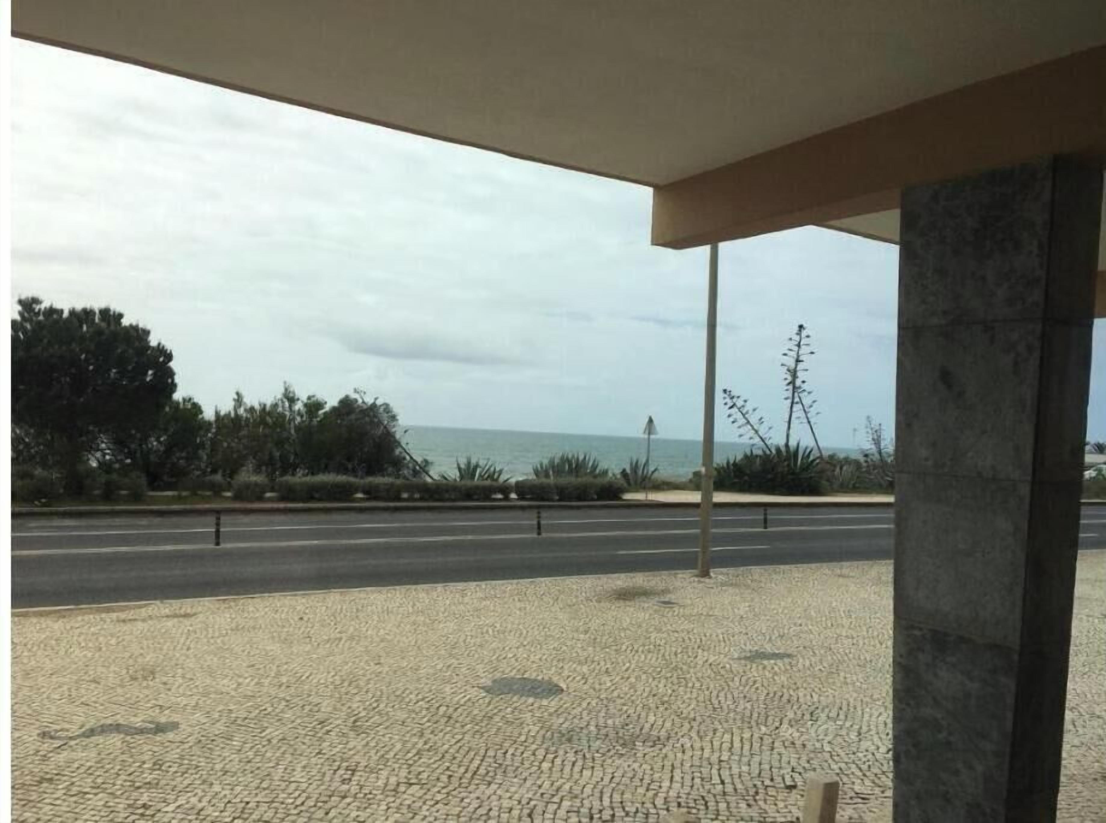 Ein überdachter Außenbereich mit gepflastertem Weg, Blick auf das Meer und einer Straße im Hintergrund.