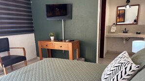 A bedroom with a bed, a desk, a chair, a TV, a mirror, and a bathroom with a sink and a lamp. - HOTEL FRANCO (El Fuerte)