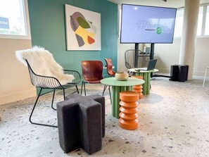 A modern interior with a green wall, a white furry chair, a colorful abstract painting, a green table with stacked stools, and a large screen displaying 'greel montpelier'. - Premiere Classe Montpellier EST Parc Expo Aeroport (Mauguio)