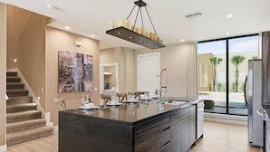 Une cuisine moderne dotée d’un grand îlot central, d’un coin repas avec des chaises, d’un escalier et d’une grande fenêtre offrant une vue dégagée.