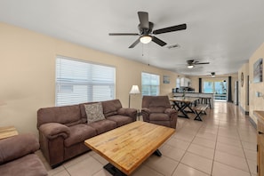 A living room with a sofa, coffee table, and ceiling fans. There is a kitchen in the background with a dining table and chairs.