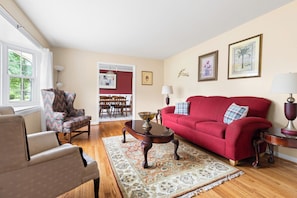 A living room with a red sofa, a patterned armchair, a wooden coffee table, and a dining area in the background. - Charming 3-bedroom house with AC in Raleigh for your stay (Raleigh)