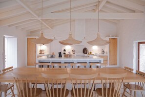 Un comedor amplio con una larga mesa de madera y sillas, una isla central con electrodomésticos de cocina, y lámparas colgantes.