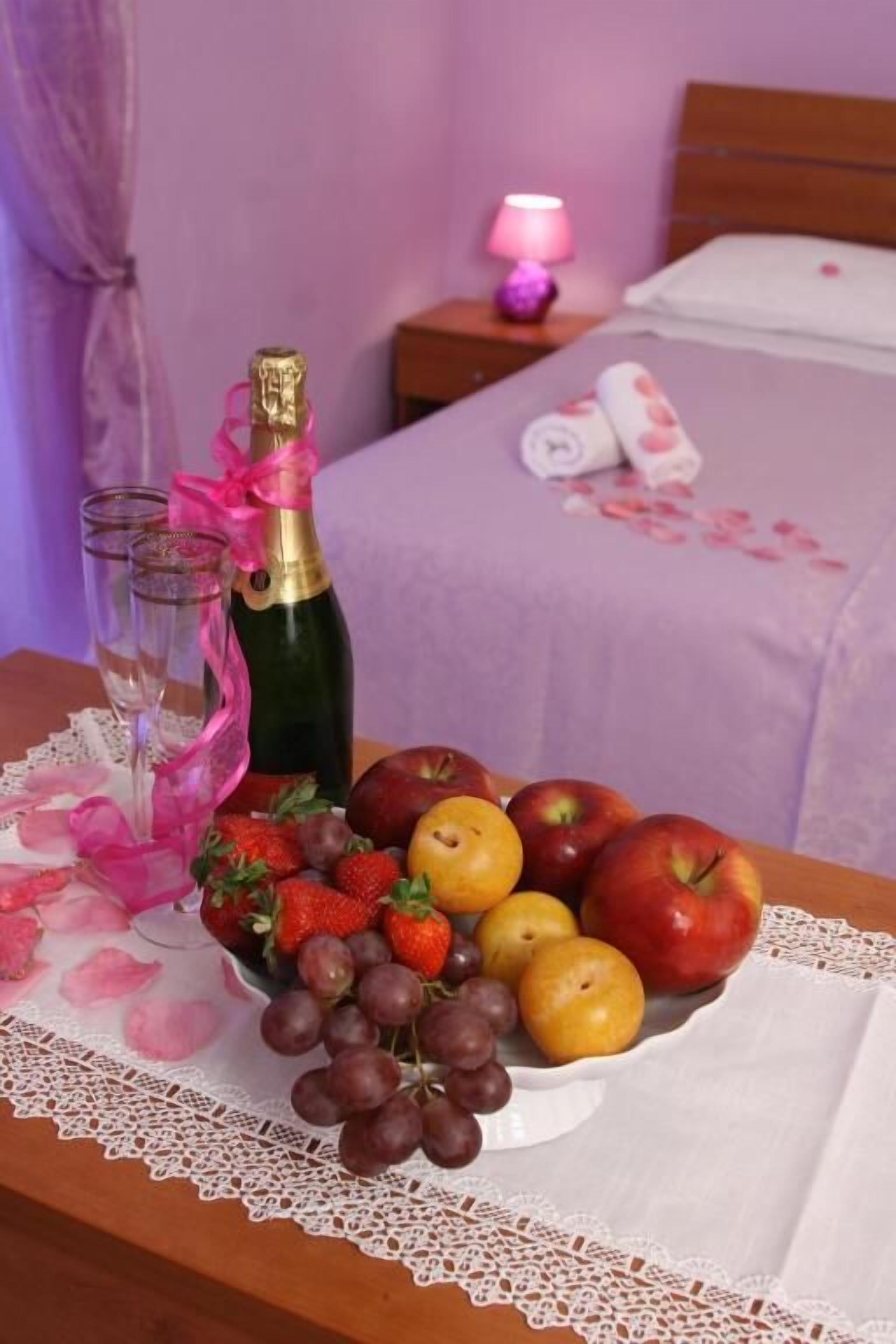 Un juego de frutas y champán dispuesto ordenadamente sobre una mesa con un mantel de encaje, una mesita de noche de madera con una lámpara, y una cama con cubrecama blanco en una habitación con paredes moradas.