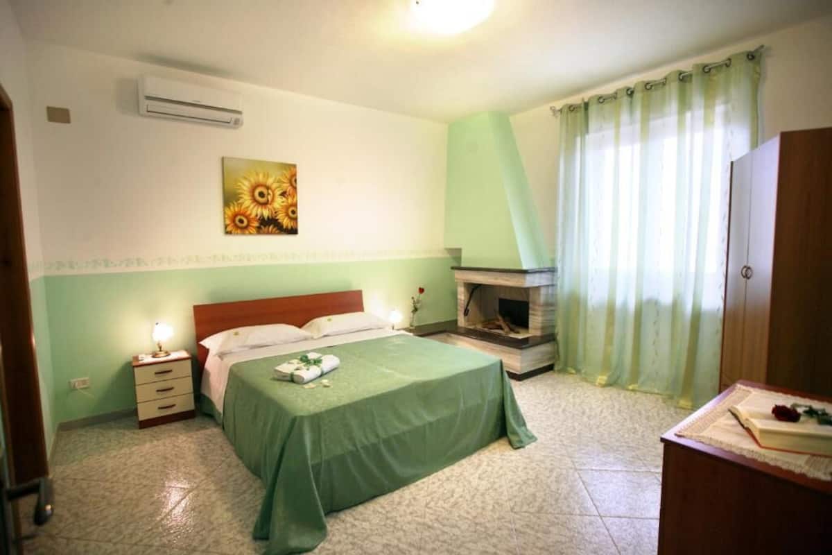 Ein Schlafzimmer mit Bett, Nachttisch, Klimaanlage an der Wand, Kamin und einem Fenster mit Vorhängen.