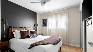 Ein Schlafzimmer mit Bett, Nachttischen, Deckenventilator, Fenster mit Vorhängen und einem an der Wand montierten Fernseher.