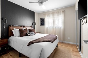 Ein Schlafzimmer mit Bett, Nachttischen, Deckenventilator, Fenster mit Vorhängen und einem an der Wand montierten Fernseher.