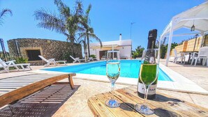 Un área junto a la piscina con sillones de descanso, una mesa de madera con una botella y copas, y un edificio de piedra al fondo.