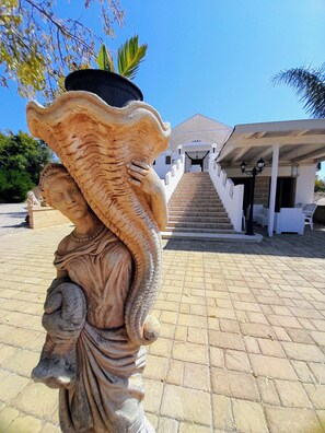 A statue with a large pot on its head, a staircase leading to a building, and outdoor seating. -  VILLA WITH TRULLO, SEA VIEW, AND EXCLUSIVE POOL (Puglia)