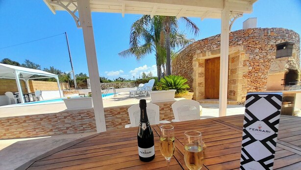 Una mesa de madera con una botella de vino, dos copas y una caja etiquetada 'FERRARI' en un espacio al aire libre techado, con un edificio de piedra y una piscina al fondo.