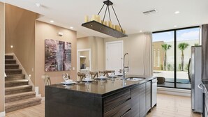 Una cucina moderna con una grande isola, un'area pranzo con sedie, una scala e vista su palme all'esterno.