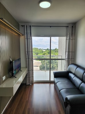 Ein modernes Wohnzimmer mit großem Fenster, einer schwarzen Ledersofa und einem Flachbildfernseher auf einem weißen Lowboard.