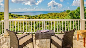 Un balcón con mobiliario de mimbre, una mesa y vistas al mar y vegetación.