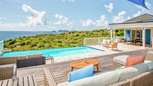 Zona junto a la piscina con mobiliario de mimbre, una terraza de madera y vistas al mar y colinas.