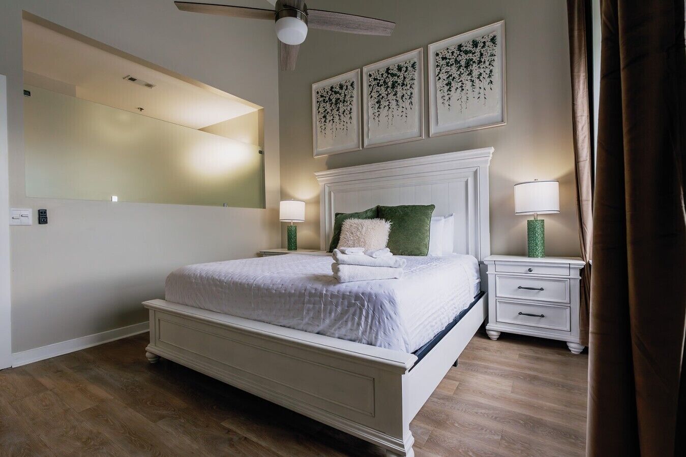 A bedroom with a bed, a nightstand, a wall-mounted ceiling fan, and framed artwork above the bed.