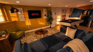 Un soggiorno con divano, tavolino, televisione che mostra 'NETFLIX', tavolo da biliardo e angolo cottura con frigorifero.