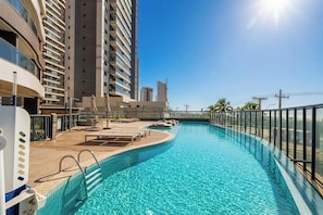 Una piscina con superficie de agua azul cristalina, rodeada por una terraza con sillas de descanso y sombrillas, y adyacente a edificios altos.
