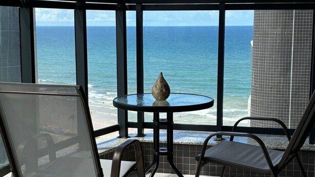 A balcony with a table and chairs, a vase, and a view of the ocean. - Beach Class Jaguaribe  (Salvador)