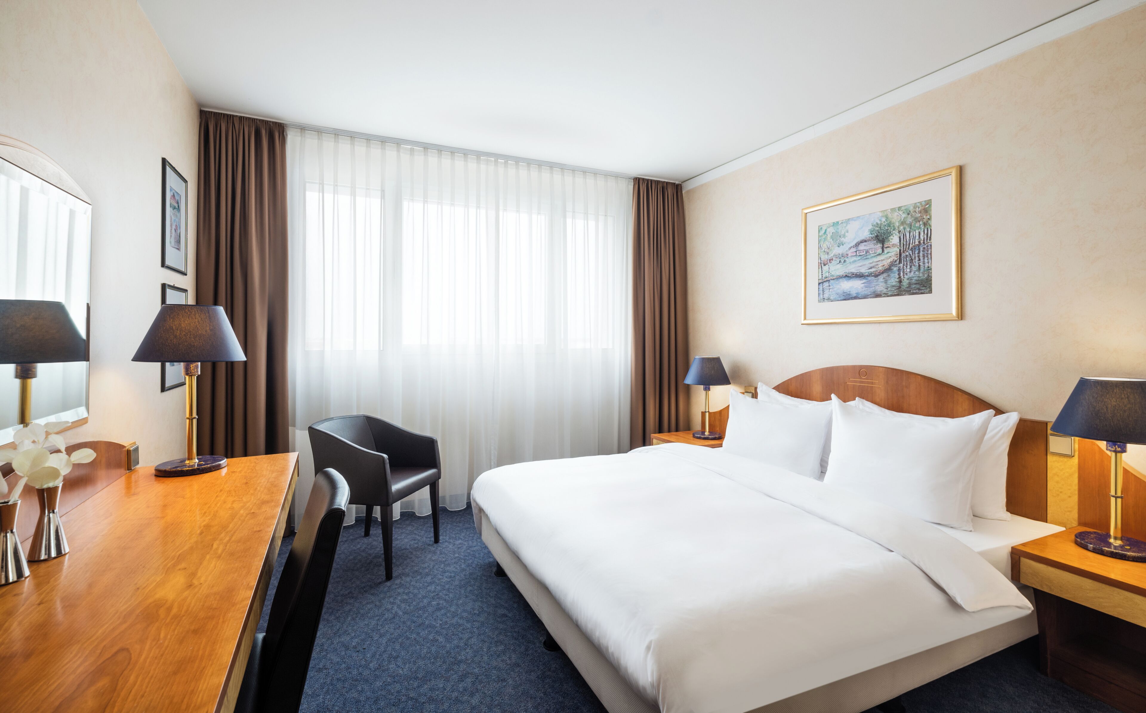 A hotel room with a bed, two bedside tables, a desk with a chair, a window with curtains, and a framed picture on the wall.
