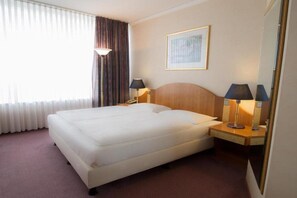 A hotel room with a large bed, two bedside tables with lamps, a framed picture on the wall, and a window with curtains. - Lindner Congress Hotel (Cottbus)