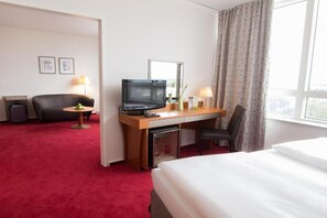 A hotel room with a bed, a desk with a television, a chair, a small table with a lamp, and a window with curtains. - Lindner Congress Hotel (Cottbus)