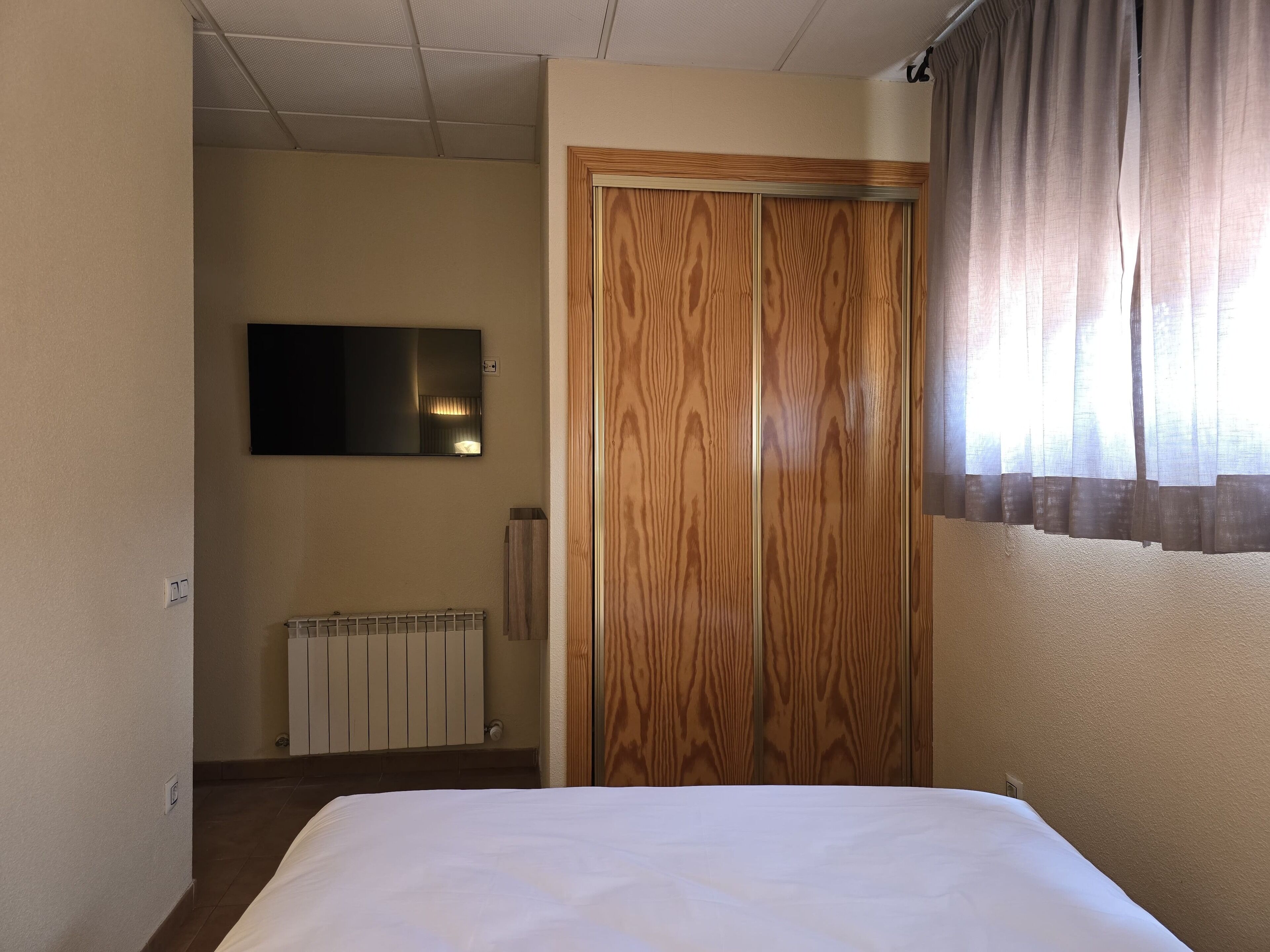 Quarto de hotel com cama, guarda-roupa de madeira, radiador e televisão na parede.