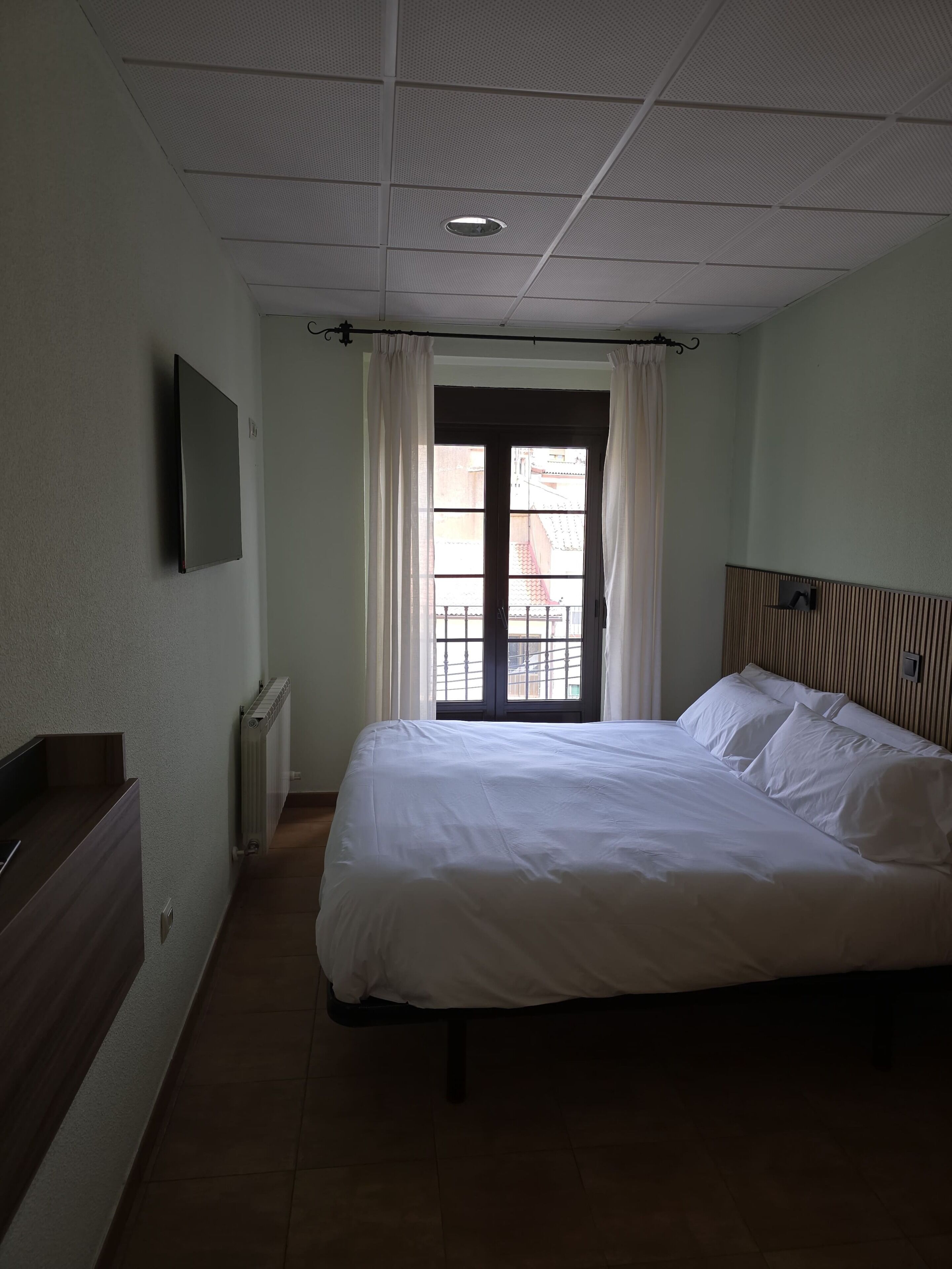 Een slaapkamer met een bed, een radiator, een raam met gordijnen, een aan de muur bevestigde televisie en een houten hoofdeinde.