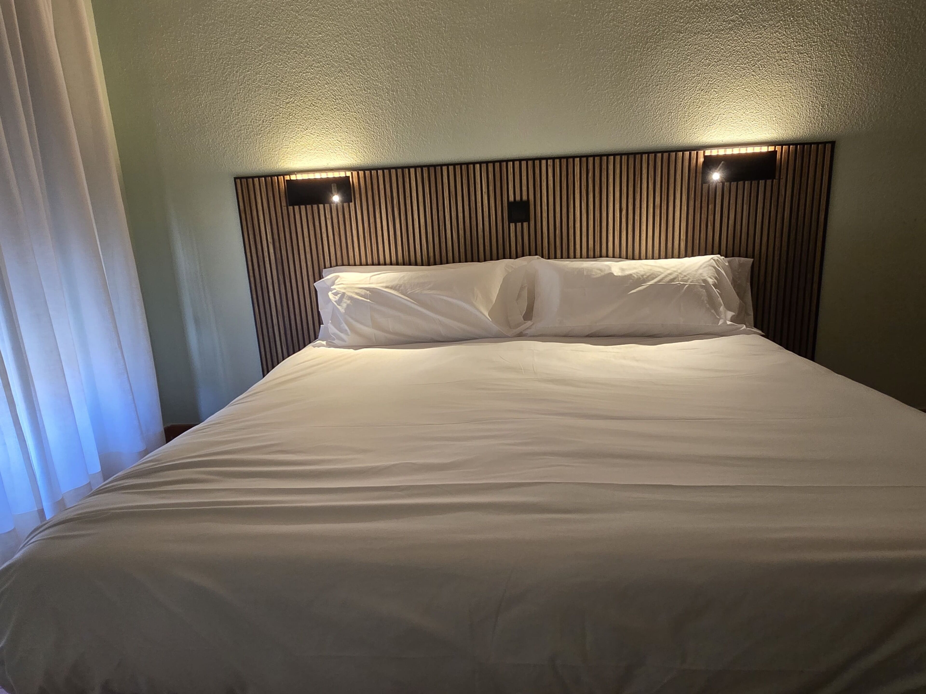 Una habitación de hotel con cama, cabecera de listones verticales y ventana con cortinas.