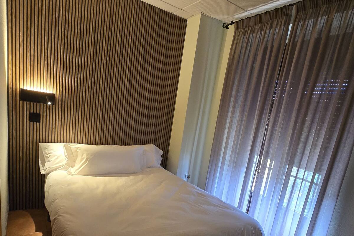 Habitación de hotel con cama, luz empotrada, ventana con cortinas y suelo de madera.