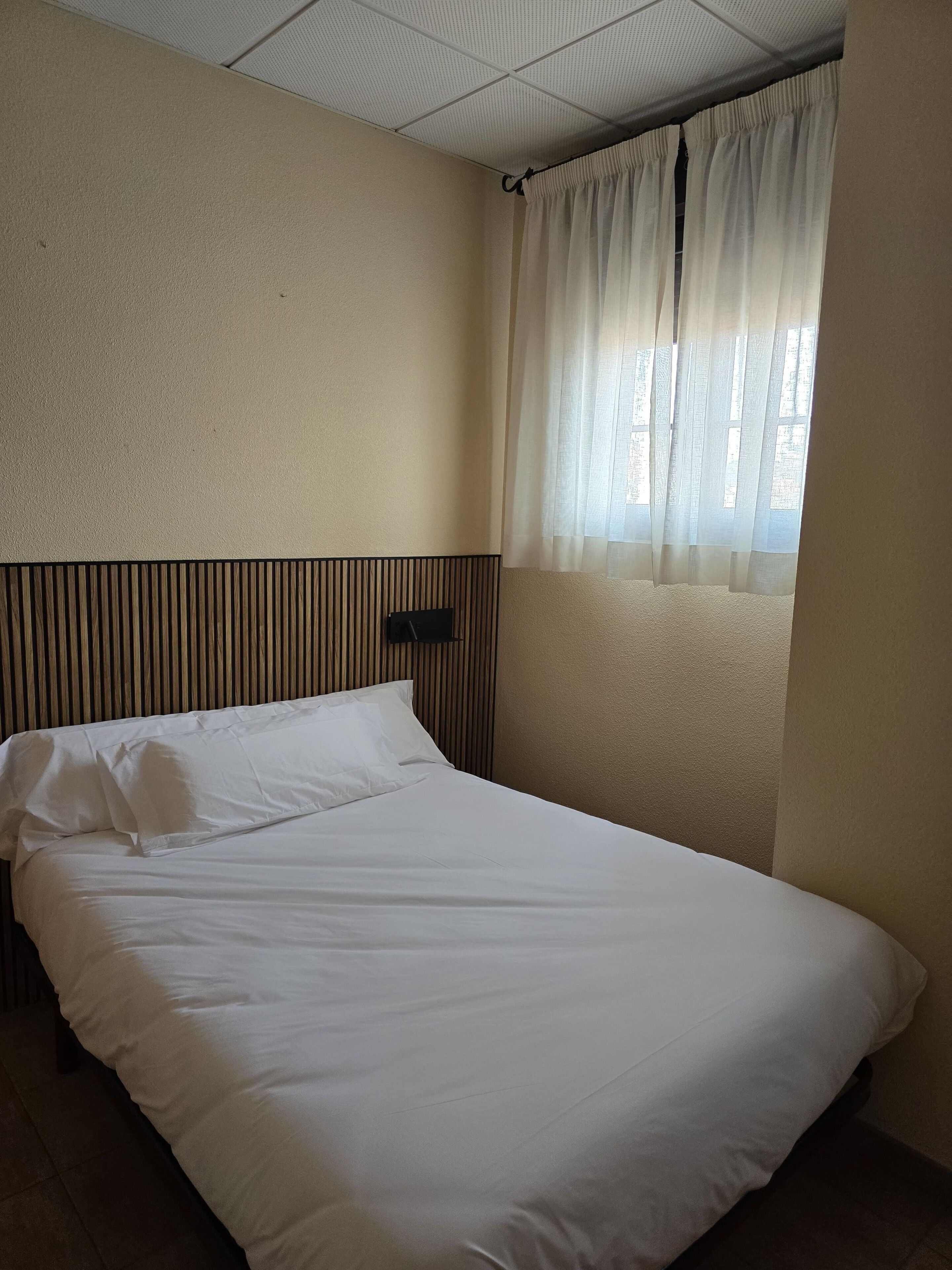 A single bed with white bedding in a room with beige walls and a window with sheer curtains.