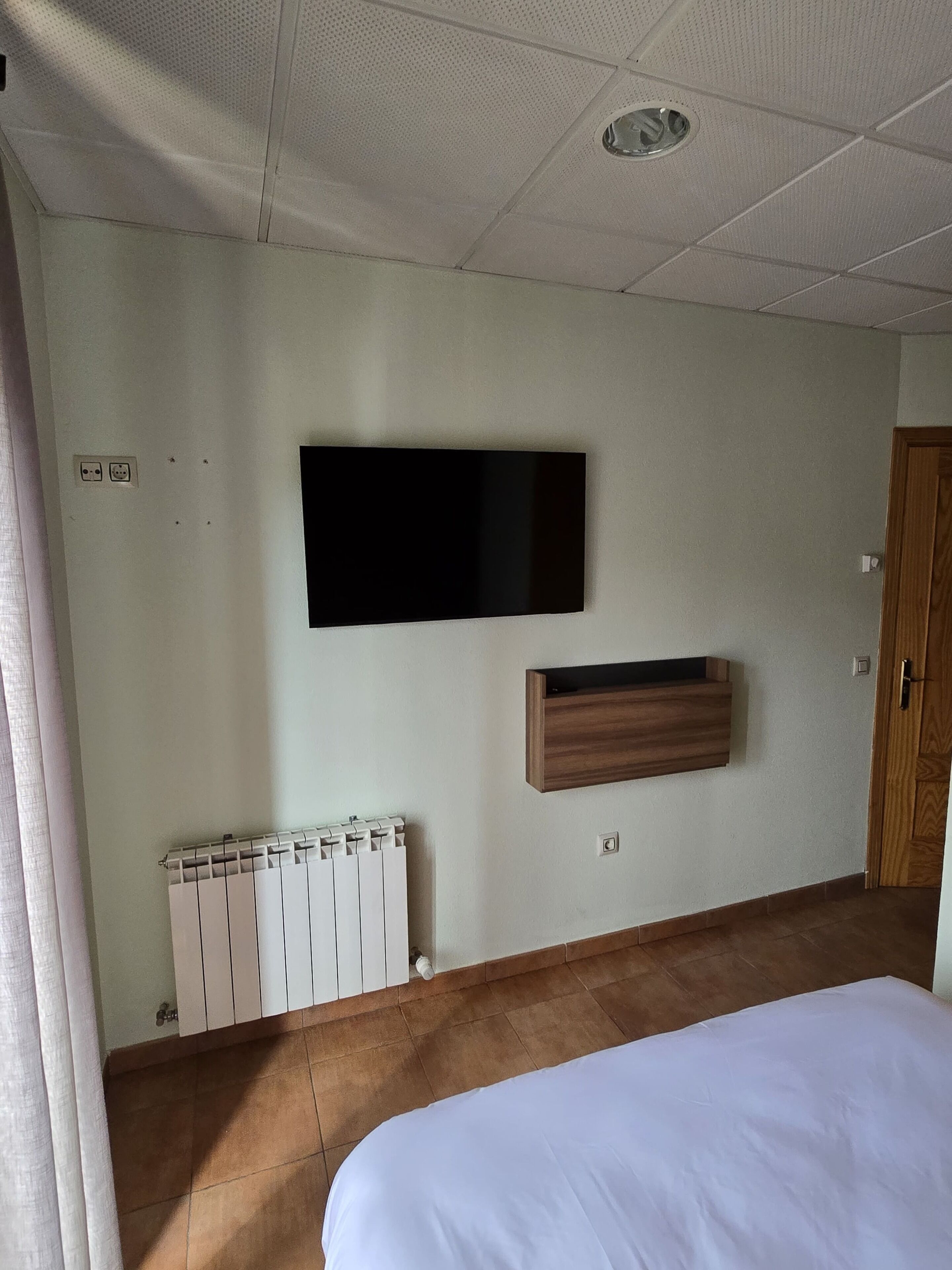 Un dormitorio con una cama, televisor montado, estante de madera, radiador y una puerta.