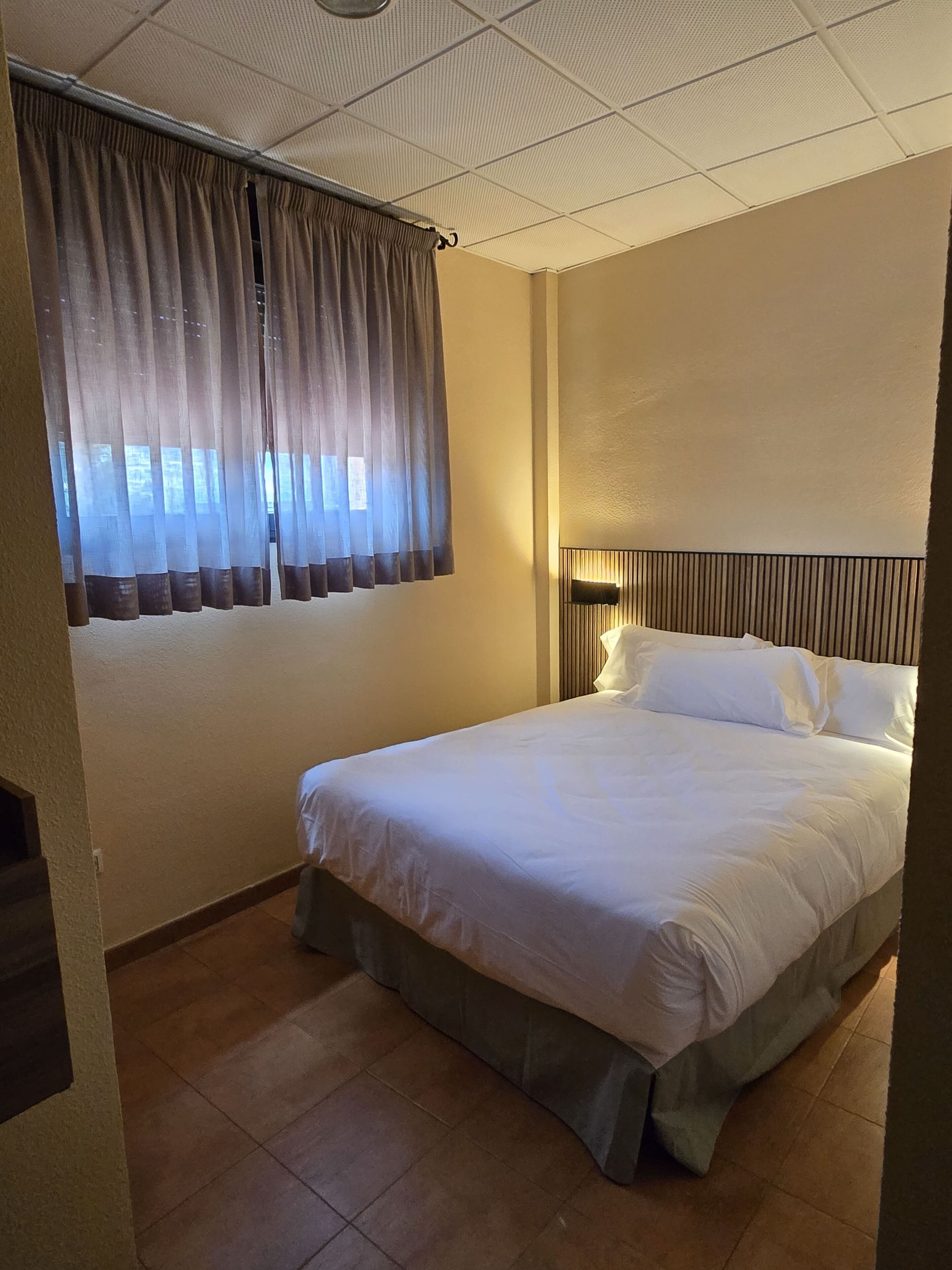 A hotel room with a bed, curtains, a headboard, a nightstand, and a wall-mounted lamp.