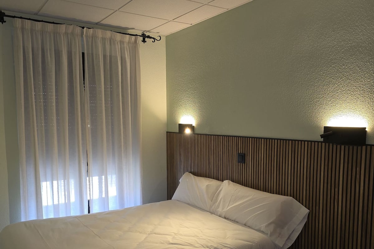 Una cama individual con cabecera, techo con luz empotrada y ventana con cortinas.