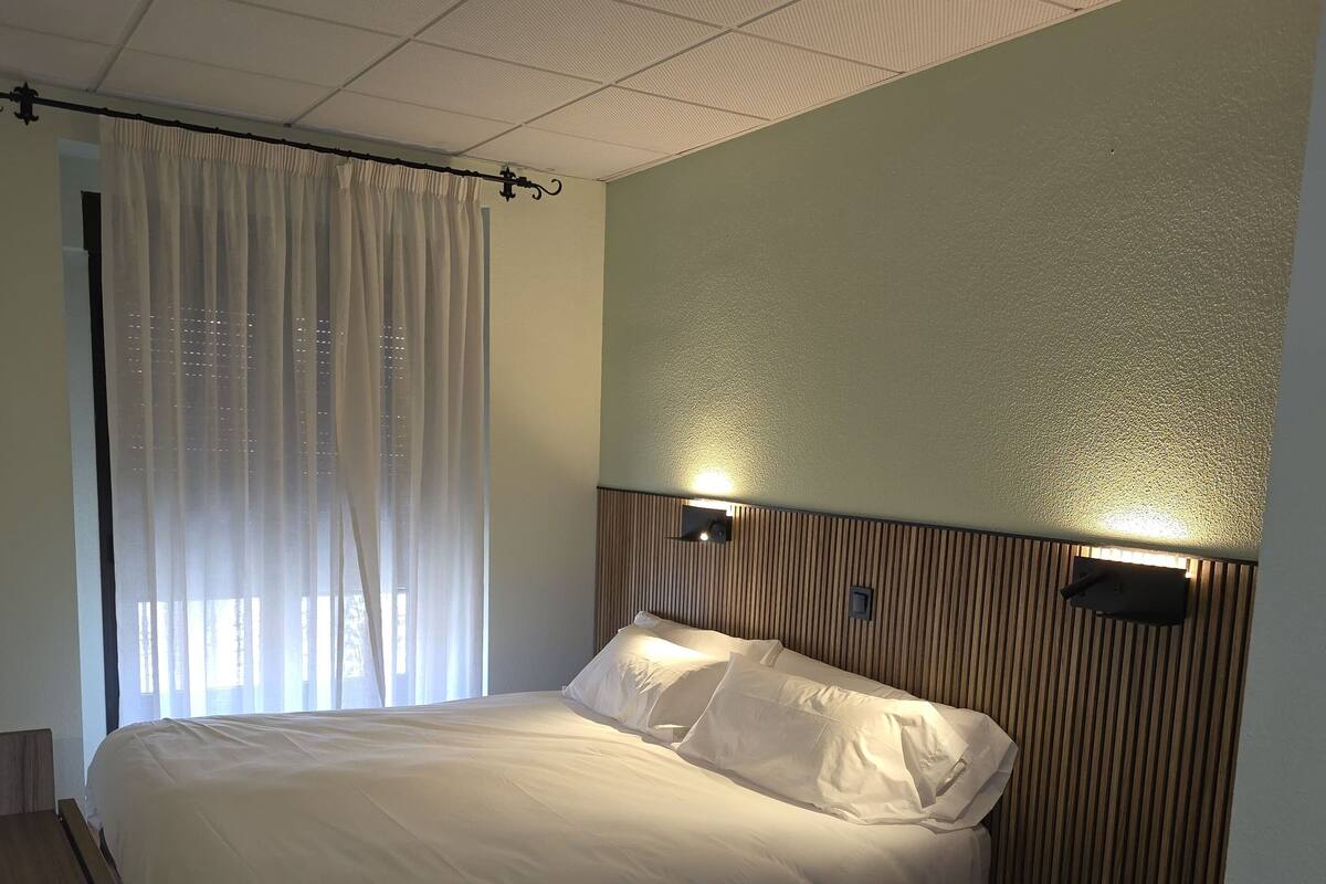 Una cama individual con ropa de cama blanca, cabecera de madera, mesita de noche con un control remoto y una lámpara de pared.