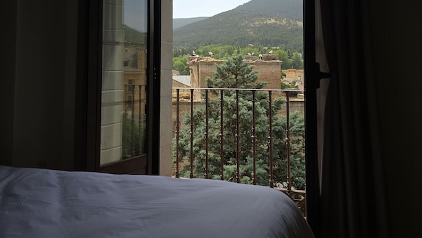 A hotel room with a bed, a window with curtains, and a view of buildings and hills. - Hostal La Cigüeña (El Espinar)