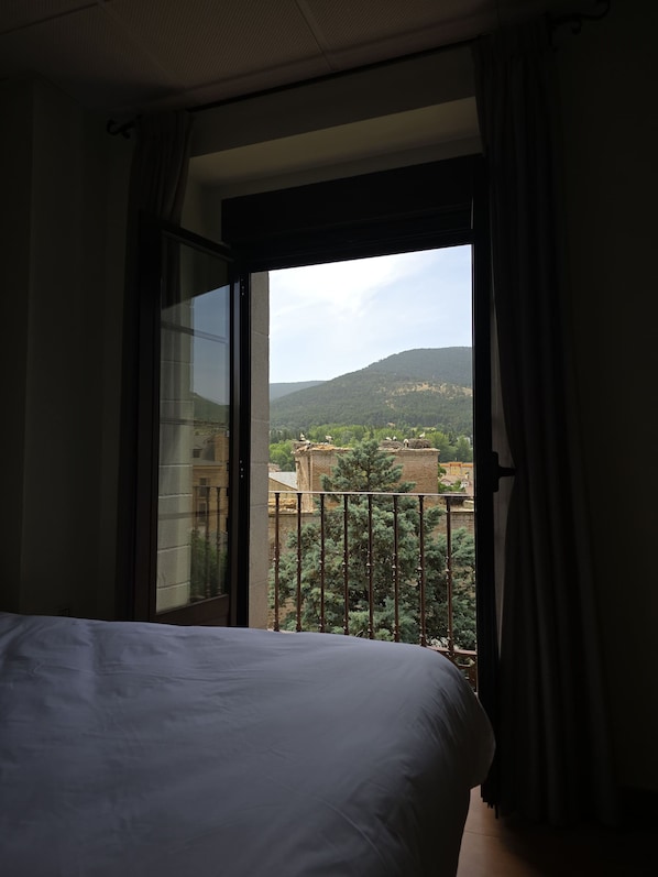 A hotel room with a bed, a window with curtains, and a view of buildings and hills.