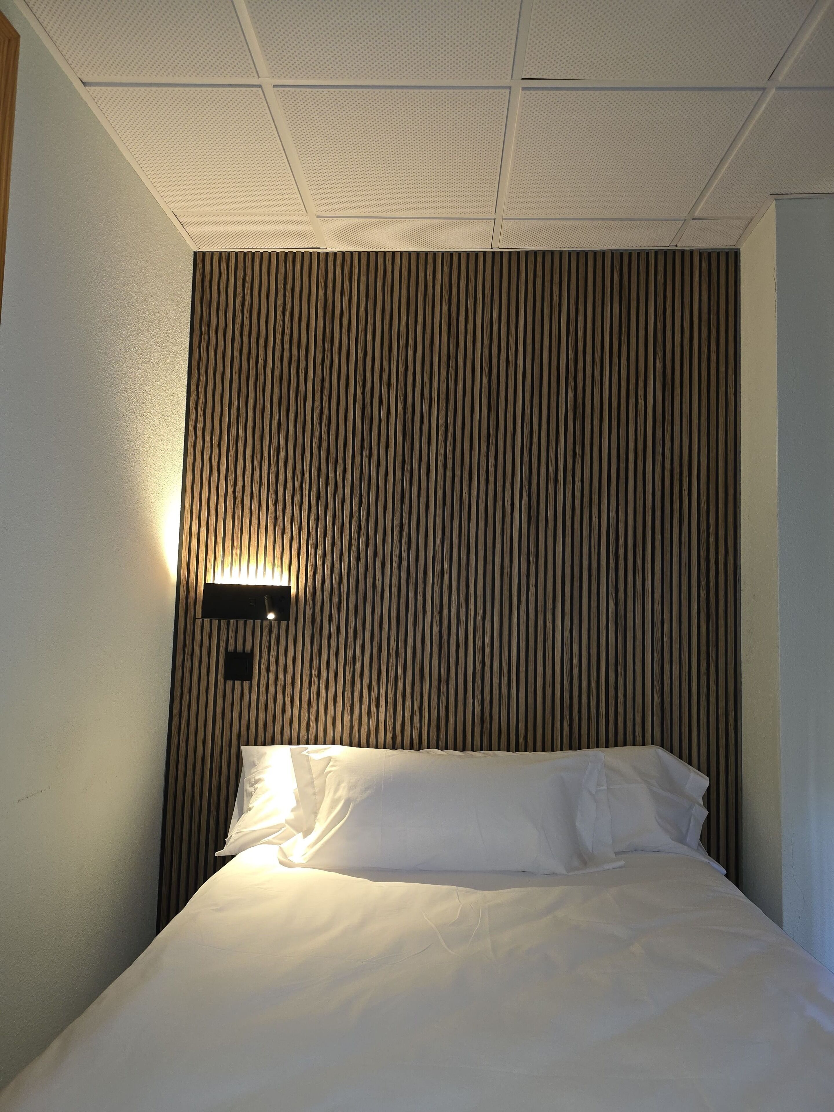 Una habitación de hotel con cama, lámpara en la pared y techo con diseño de cuadrícula.