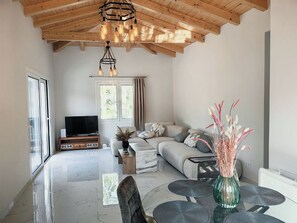 Ein geräumiges Wohnzimmer mit hoher Decke, einem großen Fenster, einer Sofagarnitur, einem Fernseher und einem Essbereich mit einem Glastisch.