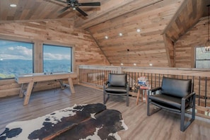 A wooden interior with a ceiling fan, a large window with a mountain view, two leather chairs, and a cowhide rug. - Indoor Pool + Panoramic Views | 4BR Sleeps 12 (Sevierville)